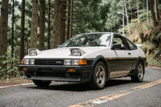 Toyota AE86 Trueno avec phares escamotables sur une route de montagne japonaise, vue trois-quarts avant mettant en valeur ses lignes angulaires distinctives